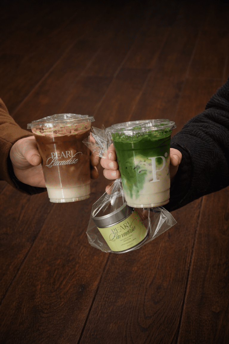 Two people toasting with Pearl Paradise branded drinks — an iced coffee and an iced matcha — over a dark wood surface. Brand lifestyle photography by Irene Villa.