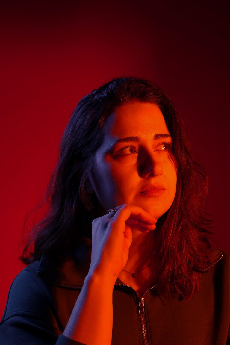 Artificial colour portrait of Nicole — warm amber and cool blue gel lighting split across a contemplative subject against a deep red backdrop. Photography by Irene Villa.